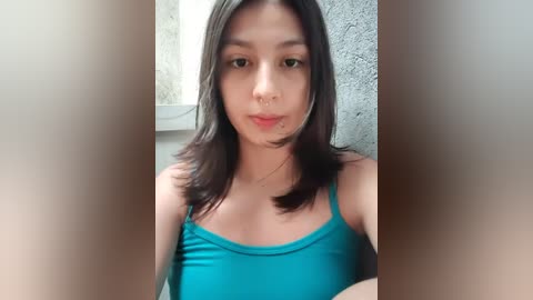 Media: A video of a young Asian woman with long black hair, wearing a teal tank top, standing against a concrete wall.