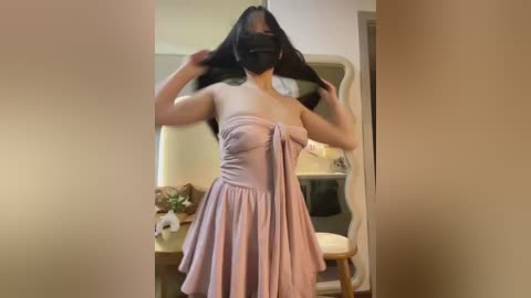 Video of an Asian woman with long black hair, wearing a pink strapless dress, black face mask, and black gloves, standing in a dimly lit room with a wooden table and a white chair.