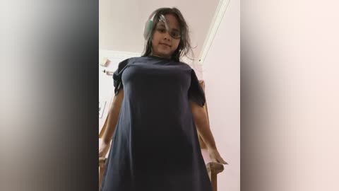 Video of a woman with dark skin and long, dark hair, wearing a black dress, standing indoors with a white door in the background.