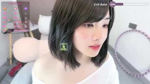 Video of a young Asian woman with straight black hair and fair skin, wearing a white off-shoulder top, sitting on a bed. Background includes a pink teddy bear and a colorful hoop.