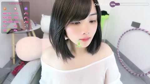 A video of a young Asian woman with straight black hair, fair skin, and a white off-shoulder top, sitting on a sofa in a minimalist room with pink balloons and a hoop.