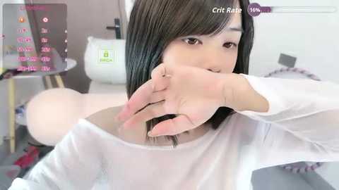 Video of an East Asian woman with straight black hair, wearing a white shirt, covering her mouth, and holding a sewing machine. Background shows a sewing table with various fabric samples.