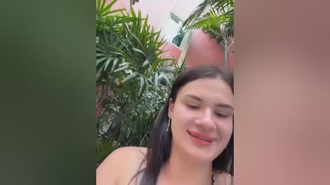 Video of a young woman with medium skin tone, straight dark hair, and a soft smile, standing in a lush green garden with vibrant foliage and a pink building in the background.