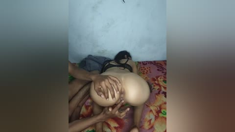 Video of a naked woman on a bed, lying on her stomach, with a man behind her, gripping her buttocks. The background is a plain white wall, and the bedspread is colorful with floral patterns.