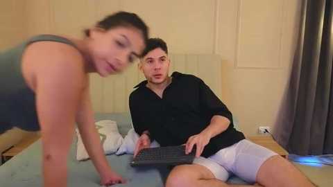 Video of a young woman with a fit physique and medium-sized breasts, wearing a gray tank top, bending over a bed. A young man with a short beard, wearing a black shirt and white shorts, sits on the bed, typing on a laptop.