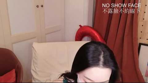 Video of a woman with long black hair, partially visible, in a cozy room with beige walls, red curtains, a brown chair, and a white cushion. Text overlay reads \"NO SHOW FACE.\