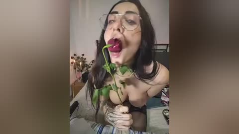 A video of a topless woman with long black hair, wearing glasses, holding a green vine with a flower, in a bedroom.