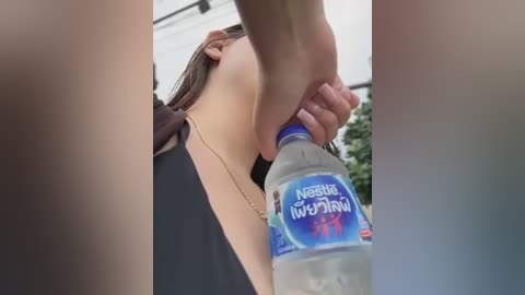 Video of a woman with long brown hair, wearing a black tank top, being fed water from a Nongshim bottle by a person's hand, blurry background.