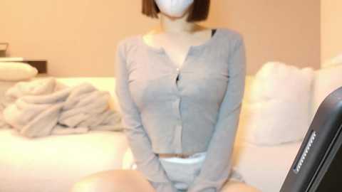 Media: Video of a woman with medium-length brown hair, wearing a grey button-up shirt and white panties, sitting on a bed with white pillows and a beige blanket.
