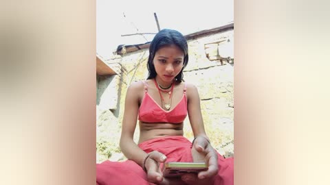 Video of a young South Asian woman with medium skin tone and long black hair, wearing a red saree, sitting on a concrete floor, holding a phone, in a rural setting with a dilapidated building in the background.