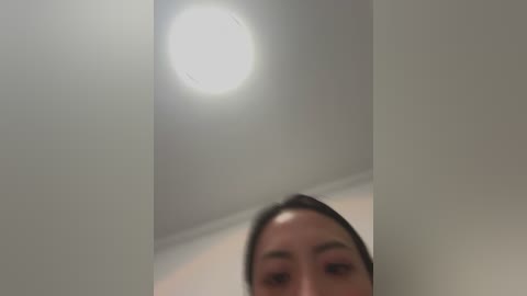 A video featuring a close-up of a woman's face with light brown skin and dark hair, partially obscured by a white wall. A bright, circular light source is visible in the background.