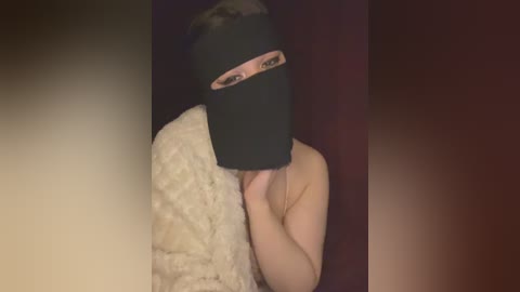 A video of a person wearing a black mask and a white, fluffy blanket, with a blurred background. The person's face is partially obscured.