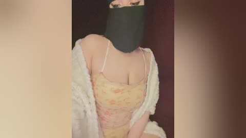 Video of a light-skinned woman with a black mask, wearing a floral-patterned yellow top, and a white shawl, against a dark background.