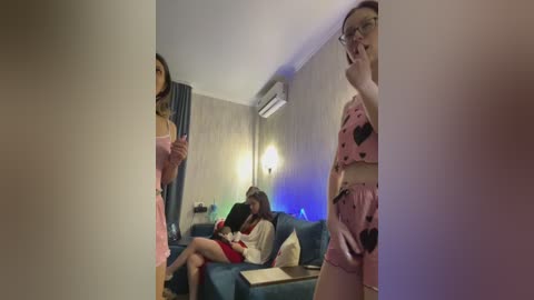 Video of a dimly lit, cozy living room with a blue sofa, featuring four people: two women in pink dresses, one man in a white shirt, and a woman with a phone.