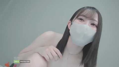 Video of a naked, fair-skinned woman with long brown hair, wearing a white face mask, in a neutral, grey background.