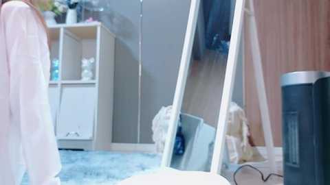 Video of a cozy bedroom with a tall mirror, white dresser, wooden wardrobe, and a blue carpet. A person in a light pink robe stands near the mirror, reflecting the room's soft lighting.