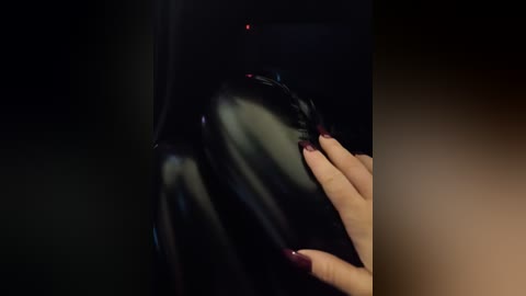 A video of a person's hand with dark red nail polish gently touching a shiny, black latex mask. The background is dark and blurred, focusing attention on the smooth, reflective texture of the mask.