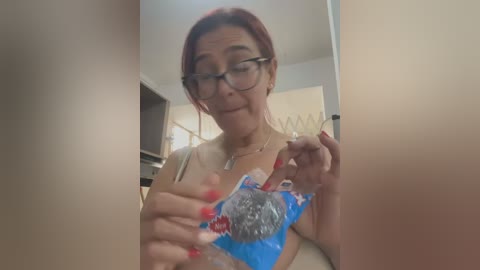 A video of a middle-aged woman with red hair and glasses, wearing a blue bra, holding a pack of cookies in a cluttered kitchen.