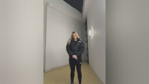 Video of a blonde woman in a black puffer jacket and leggings standing in a dimly lit, narrow hallway with beige walls and a single light fixture on the right.