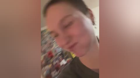 Blurry video of a young, fair-skinned woman with short, dark hair, wearing a dark top. The background is indistinct, possibly a wall with colorful posters. The image appears to be taken with a shaky camera, giving a sense of motion.