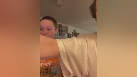 Video of a fair-skinned, short-haired boy wearing an orange shirt with white patterns, looking at a mirror; background shows a wall with various posters and a ceiling light.