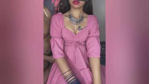Video of a woman with long dark hair, wearing a pink dress and black blindfold, silver necklace, and black bracelets, seated on a couch in a dimly lit room.