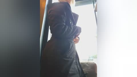 Video of a woman with light skin and blonde hair, wearing a black trench coat, standing inside a dark, narrow doorway, with a blurry, sunlit background.