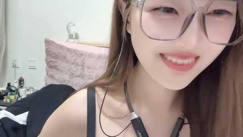 Media: A video of an Asian woman with fair skin, long brown hair, and glasses, smiling, wearing a black tank top, sitting on a bed with a pink blanket.