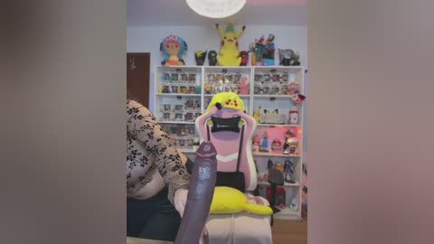 Media: Video of a person wearing a Pikachu costume in a room with Pikachu plushies, a pink blanket, and shelves filled with toys and books.