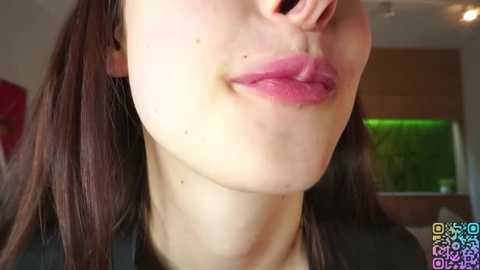 Video of a close-up of a woman's face, showing her fair skin, pink lips, and brown hair, with a blurred background featuring a window and green foliage.