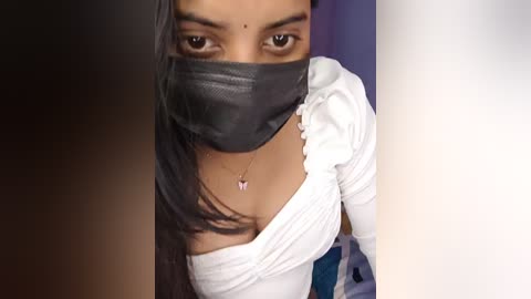 Video of a young woman with medium brown skin, long black hair, and dark eyes, wearing a black face mask and a white off-shoulder top with ruffles, set against a blurred purple and white background.