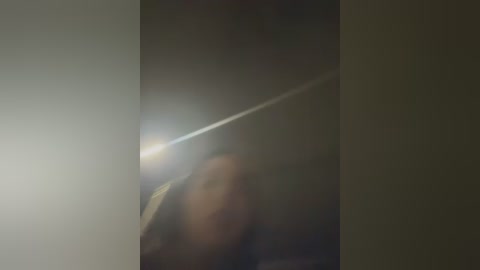 A blurry video of a person, possibly a woman, in a dark, dimly lit room. The image is grainy, with a bright, long streak of light cutting across the frame, suggesting a camera flash or a moving object.