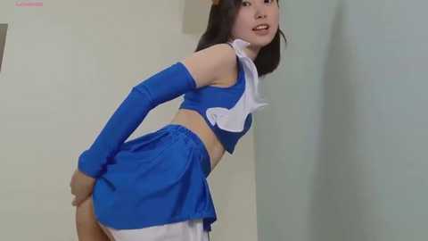 Video of an Asian woman in a blue cheerleader outfit with white accessories, bending over slightly, with a light green wall in the background.