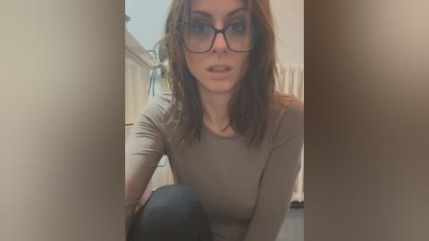 Media: Video of a Caucasian woman with shoulder-length brown hair, wearing black-framed glasses and a beige long-sleeve top, crouching in a hallway with beige walls, looking surprised.