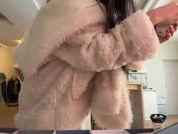 A video of a woman in a fluffy, light pink robe, bending over, revealing her thighs, in a modern, cluttered living room with a white door and a pile of clothes.
