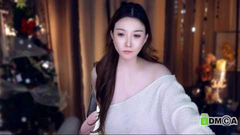 Media: Video of a young Asian woman with long, dark hair, wearing a white off-shoulder sweater, standing indoors near a Christmas tree and festive decorations.