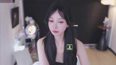 Video of a young East Asian woman with long black hair and fair skin, wearing a black top, seated in a modern studio with black backdrop, lighting equipment, and framed art.