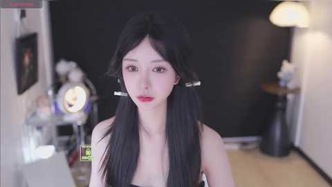 Video of a young Asian woman with long black hair in pigtails, wearing a black dress, in a dimly lit room with a black background, lamps, and a cluttered desk.