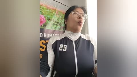 Video of an East Asian woman with glasses and long black hair, wearing a black and white athletic jacket with number 23, standing in front of a colorful wall poster.