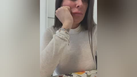 Media: Video of a woman with long, straight brown hair, wearing a beige long-sleeve top, resting her chin on her hand, partially obscuring her face, in a bathroom with white tiles.