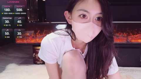 Video of an Asian woman with long black hair, wearing a white T-shirt and face mask, kneeling on a white bed in a dimly lit room with a fireplace and TV screen showing game statistics.