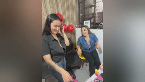 Video of two women in an office: one with long black hair and a black shirt, laughing; the other with medium-length brown hair and a blue shirt, smiling. Red balloons and papers on desks, beige curtains, and a whiteboard with text.