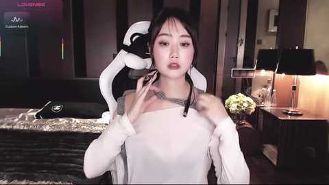 Video of an East Asian woman in a white off-shoulder dress, holding a headset, indoors with modern decor, a lamp, and a table in the background.