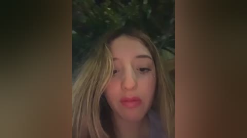 Video of a young woman with long, straight blonde hair, fair skin, and full lips, smiling slightly, set against a blurred, dark green foliage background.