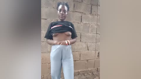 A video of a young Black woman with medium skin tone, wearing a black T-shirt lifted to reveal small, round breasts. She has short, curly hair and wears light blue pants. The background is a beige brick wall.