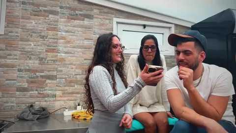 Video of three young adults, one in a grey sweater, another in a beige sweater, and the third in a white shirt and cap, sitting on a rooftop patio with a brick wall background and a grill nearby.