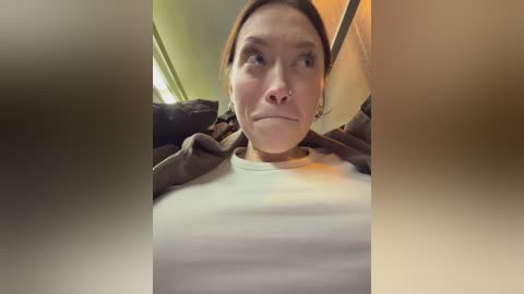 Media: Video of a woman with a nose ring, brown hair, and a white shirt, taken from an upward angle, showing a distressed expression, surrounded by blurred, dimly lit room.