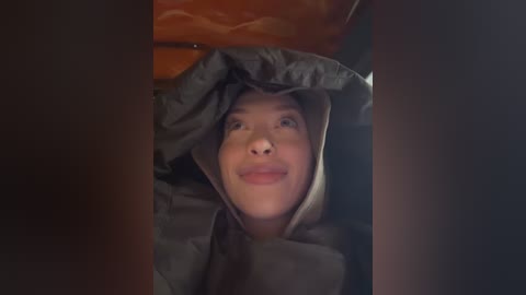 Media: A video captures a smiling woman with light skin and short black hair, wearing a dark, hooded raincoat, peeking from under a car's open hood.