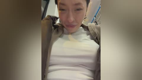 Media: Video of an Asian woman in her late 20s with short black hair, wearing a white t-shirt under a brown jacket, sitting in a car. Her face is partially obscured by the camera's focus on the steering wheel.