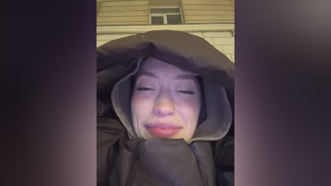 Media: A video of a person in a black hooded jacket, partially covering their face, standing in front of a beige house with a small window.